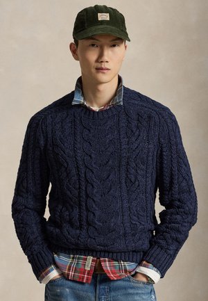 Jeune homme portant un pull torsadé bleu foncé sur une chemise à carreaux, associé à un jean bleu et une casquette en velours côtelé vert foncé.