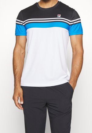 Mann trägt kurzärmliges T-Shirt mit farblich abgesetzten schwarzen, weißen und blauen horizontalen Streifen und schwarze Hose, steht mit einer Hand in der Tasche.