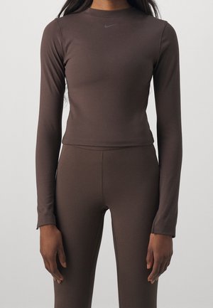 Kvinde iført tætsiddende mørkebrun langærmet top og matchende leggings med subtilt logo på brystet, stående mod en ensfarvet baggrund.