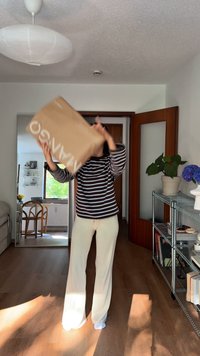 Osoba v pruhovanom hnedobielom tričku s dlhým rukávom a krémových širších nohaviciach zdvíha hnedý balík s bielym logom v jasne osvetlenej miestnosti.