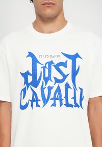 White cotton t-shirt featuring bold, textured blue lettering "JUST CAVALI" and "FIRED EARTH" in a smaller font above, round neck design.