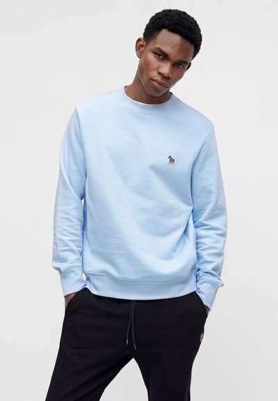 Jeune homme portant un sweat-shirt bleu clair avec un petit logo zèbre sur la poitrine et un pantalon noir, les mains dans les poches, debout sur un fond uni.