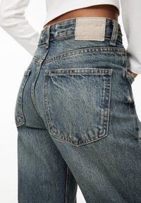 Jeans de mezclilla azul de cintura alta con un lavado desgastado, que cuentan con bolsillos traseros, detalles de costura y un parche de cuero beige claro en la cinturilla.