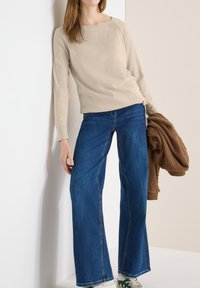 Personne portant un haut beige à manches longues et un jean bleu à jambes larges, tenant une veste marron texturée, debout contre un mur bicolore.