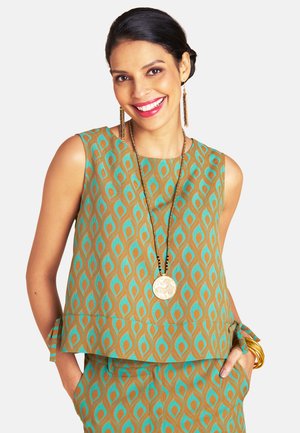 Frau lächelt, trägt ärmelloses türkis-braunes gemustertes Oberteil mit passender Hose, goldenen Schmuck und lange Anhängerhalskette, Hände in den Taschen.