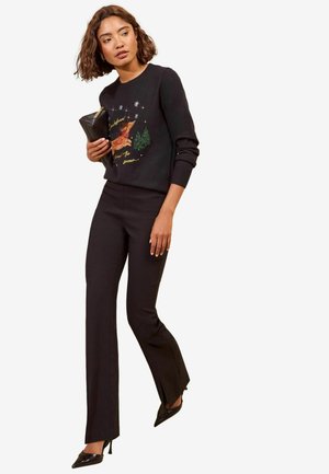 Frau trägt einen schwarzen Pullover mit Rentier und Weihnachtsbäumen, schwarze Hose, schwarze High Heels und hält eine schwarze Clutch.