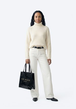 Hosszú sötét hajú nő, krémszínű garbót és fehér nadrágot visel, kezében egy fekete Ted Baker London válltáskával.