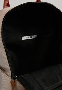Vaaleanruskea tikkikuvioinen reppu, jossa on musta vuori, hopeinen vetoketju ja "PIECES" -merkki ommeltuna sisäpuolelle. Näkyvissä ruskea nahkakoriste.