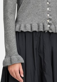 Grauer Strickcardigan mit Rüschensaum und Glockenärmeln. Sechs Knöpfe vorne, kombiniert mit einem dunklen, strukturierten Rock.