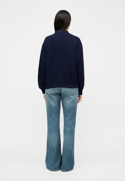 Γυναίκα με μακριά σκούρα μαλλιά που φοράει σκούρο μπλε πουλόβερ και ανοιχτό μπλε τζιν με καμπάνες, όρθια με την πλάτη προς το μέρος μας σε απλό λευκό φόντο.