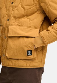 Gequiltede pufferjack in mosterdgeel met twee voorzakken, drukknopen en een zwart Timberland logo patch op de linkerzak.