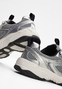 Scarpe da ginnastica argento con dettagli in rete, suola intermedia bianca e suola nera; hanno un tirante e lacci rotondi.