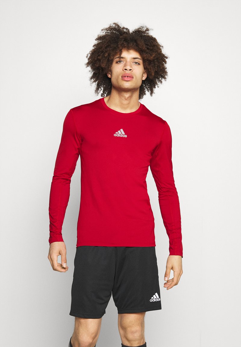 Rotes Langarmshirt mit Rundhalsausschnitt und silbernem Adidas-Logo. Kombiniert mit schwarzen Sportshorts mit weißem Adidas-Logo.
