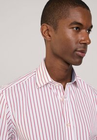 Chemise blanche avec des rayures verticales rouges, col boutonné, et une bordure intérieure de col bleu clair. Tissu en coton lisse, design ajusté.