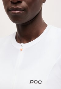 Κοντινό πλάνο ατόμου που φοράει λευκό ποδηλατικό μπλουζάκι POC με μικρό φερμουάρ μπροστά και στρογγυλή λαιμόκοψη, εμφανίζοντας το κάτω μέρος του προσώπου και το λαιμό.