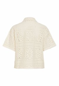 Blouse crème à manches courtes avec col, présentant un motif découpé ajouré géométrique et floral sur toute la surface.