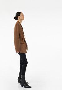 Blazer marrón con cinturilla, usado sobre jeans skinny negros, combinado con botas negras hasta el tobillo con tacón grueso.