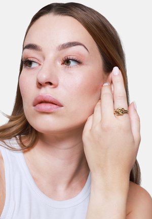 Goldfarbener Ring mit verflochtenen Bändern und einer strukturierten Oberfläche. Das Model berührt sanft ihr Gesicht und präsentiert das Design des Rings.