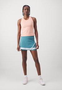 Top fără mâneci în nuanță piersică deschisă, cu un design ajustat și logo. Fustă închisă la culoare, cu un model tip wrap, suprapusă peste pantaloni scurți ajustați și decorată cu un tiv alb. Pantofi atletici albi.