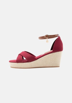 Sandalia de cuña roja con tiras cruzadas, suela de plataforma tejida y correa marrón en el tobillo con un pequeño charm dorado.