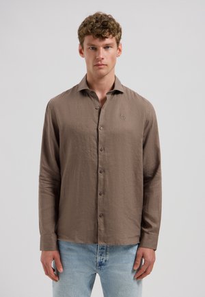 Junger Mann mit lockigem Haar, der ein braunes Hemd mit langen Ärmeln und hellblaue Jeans trägt, vor einem schlichten hellen Hintergrund stehend.