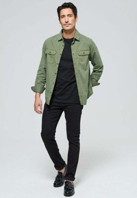 Chemise en coton verte sur un t-shirt noir, accompagnée de jeans noirs et de mocassins noirs. La chemise comporte deux poches poitrine et des manches retroussées.