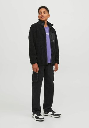 Giacca in pile nera con colletto alto, tasca con zip sul petto, abbinata a una t-shirt viola, pantaloni cargo neri e sneakers con dettagli bianchi.
