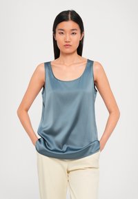 Woman with straight black hair wearing a sleeveless shiny blue top and light beige pants, standing with hands in pockets against plain background.
