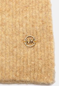 Nærbillede af struktureret beige strikket stof med et lille rundt guld metal MK-logo fastgjort i midten.