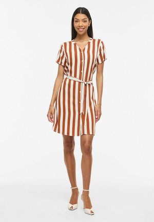 Kobieta ubrana w sukienkę z krótkim rękawem zapinaną na guziki, z pionowymi paskami w kolorze rdzy i białym, związaną paskiem w talii, stojąca i uśmiechająca się, w białych szpilkach.