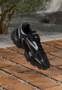 Zapato deportivo negro con parte superior de malla, acentos plateados y suela de goma. Presenta un diseño de cordones y soporte acolchado para el tobillo.