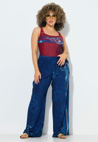 Kvinde med krøllet hår iført gule solbriller, en rød ærmeløs top og løse blå bukser med mønster samt gyldne sandaler, stående foran en hvid væg.