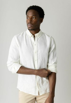 Mand med kort krøllet hår, der ruller ærmet op på en hvid knappet skjorte, kigger til siden mod en ensartet lys baggrund.