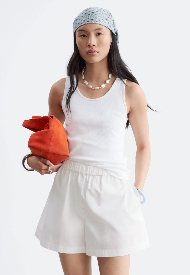 Femme en débardeur blanc et short, portant un foulard à motifs et un collier de coquillages, tenant un sac orange et portant des bracelets.