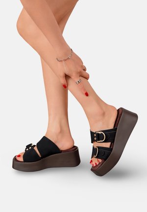 Jambes portant des sandales à plateforme noires avec boucles dorées, ongles de pieds peints en rouge, et une main touchant la cheville droite ornée de bagues en argent et d'un bracelet.