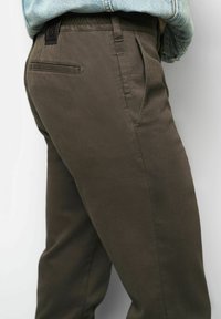 Donkergroene broek met een gladde textuur, voorzien van een enkele achterzak, plooien en een subtiele taillebandlabel voor merkaanduiding.