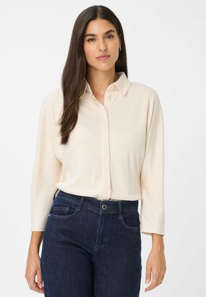 Woman with long dark hair wearing a cream button-up blouse and dark blue jeans, standing against a plain white background.