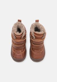 Une paire de bottes brunes pour enfants avec doublure en fourrure et deux bandes velcro, vues d'en haut sur un fond blanc.