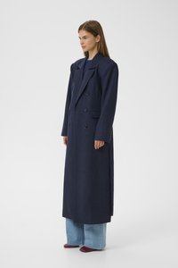 Navy blue long wool coat with a double-breasted design, notched lapels, front pockets, and side slits. Paired with wide-leg jeans and red shoes.