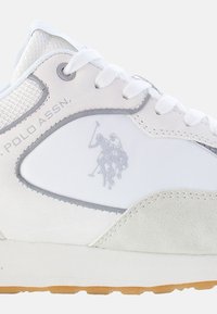 Sneaker bianca con un mix di pelle e camoscio, dettagli grigi e un logo di un giocatore di polo discreto sul lato. Parte superiore in rete tessile.