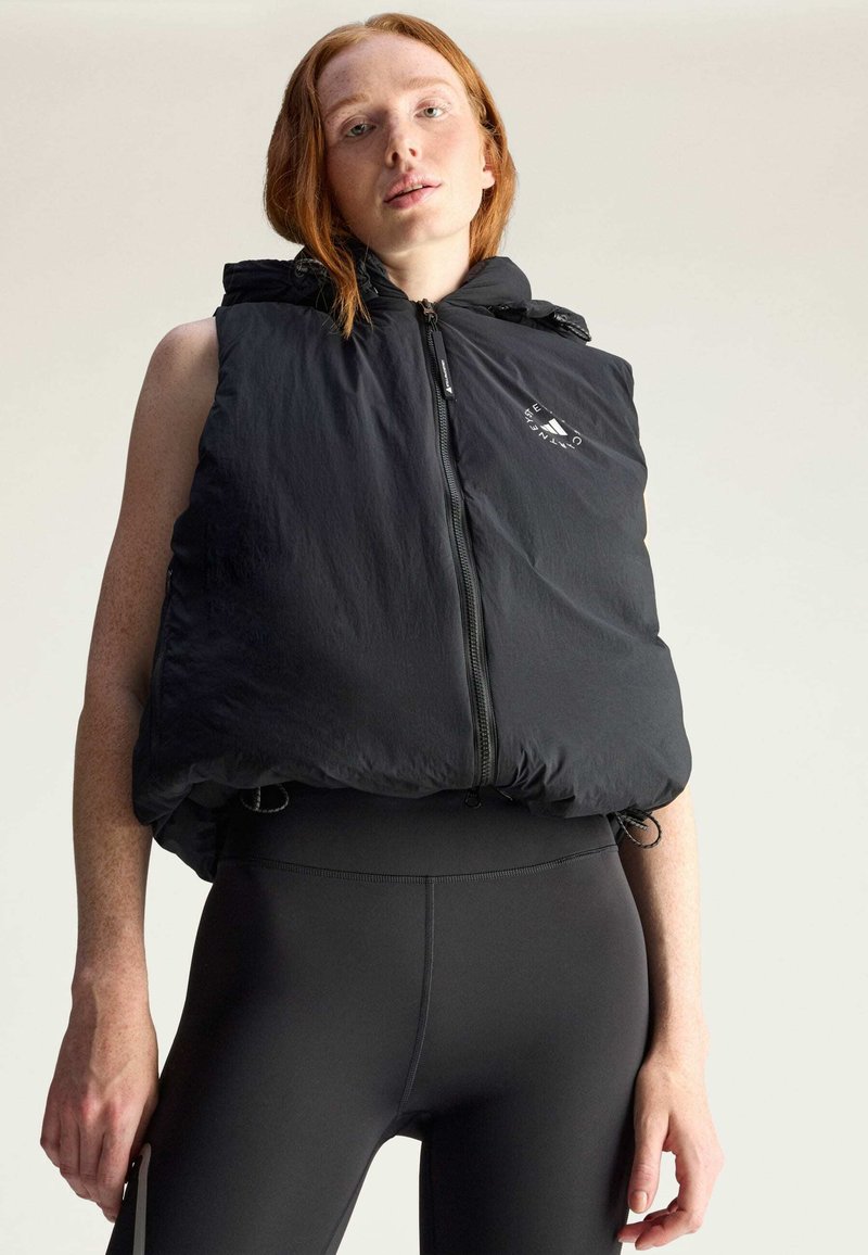Zwarte gewatteerde vest met ritssluiting en capuchon, voorzien van een subtiele logo op de borst. Gepaard met strakke zwarte leggings.