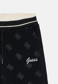 Melni treniņbikses ar ribotu jostu, ar izbordētu "Guess" logo un smalku visā virsmā redzamu zīmola iniciāļu rakstu.