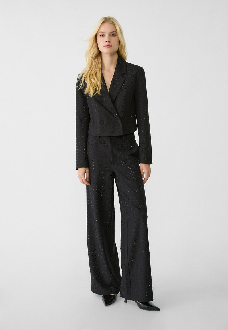 Blazer corto negro ajustado con botones de doble botonadura y pantalones de pierna ancha a juego, ambos con sutiles adornos y una textura suave.