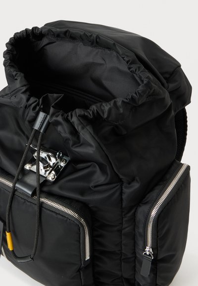 Sac à dos en nylon noir avec ouverture supérieure à cordon, poches avant et latérales zippées, et fermeture à boucle argentée.