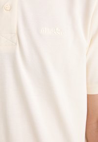 Polo shirt color crema realizzato in tessuto texturizzato. Presenta un colletto con chiusura a tre bottoni e un logo in rilievo sul petto. Maniche corte.