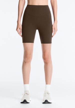 Stehende Person trägt braune, hoch taillierte Biker-Shorts, weiße Sportsocken und cremeweiße Sportschuhe vor einfachem Hintergrund.