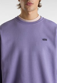 Sudadera lavanda con cuello redondo, hecha de algodón. Presenta un pequeño logo negro "VANS" en el lado izquierdo del pecho. Textura suave.