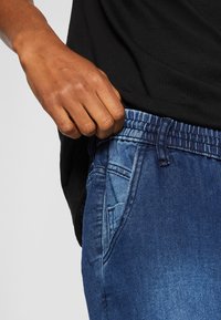 Ręka dostosowująca elastyczny pas niebieskich dżinsów noszonych z czarną koszulą, pokazująca szczegóły przedniej kieszeni.