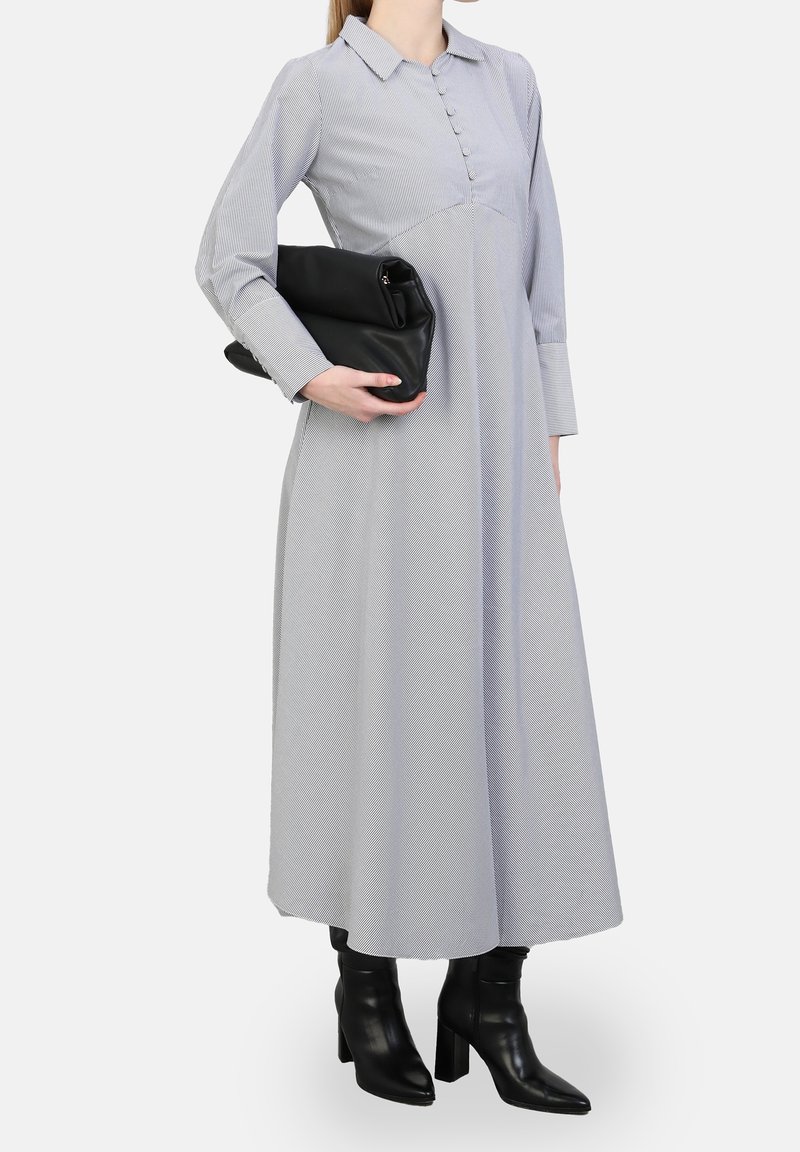 Femme portant une robe grise à manches longues, longueur cheville, tenant une pochette noire, associée à des bottines noires à talons.