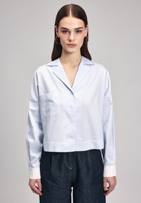 Camicia a righe in blu chiaro e bianco, maniche lunghe, design cropped, chiusura a bottoni, taschino sul petto, texture liscia, abbinata a pantaloni in denim scuri.